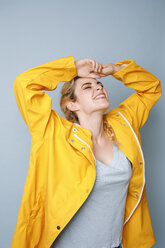 Happy young woman wearing yellow rain coat in front of blue background - GRSF00055