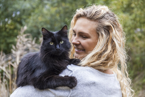 Porträt einer schwarzen Katze auf der Schulter einer lächelnden jungen Frau - TCF06064