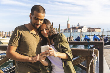 Italien, Venedig, verliebtes junges Paar mit Handy und Gondelbooten im Hintergrund - WPEF01254