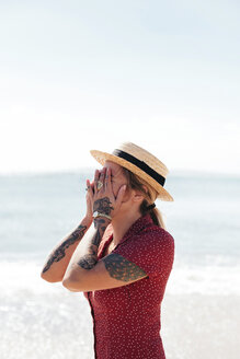Young woman with tattoos on hand and arms covering her face in front of the sea - LOTF00018