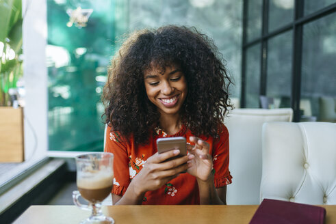 Porträt einer lächelnden jungen Frau in einem Café, die ein Mobiltelefon benutzt - KIJF02138