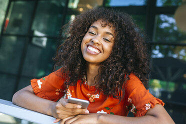 Portrait of laughing young woman with smartphone leaning on window sill of a coffee shop - KIJF02149