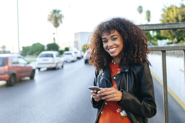 Portrait of smiling young woman with smartphone standing at roadside - KIJF02167