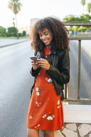 Porträt einer lächelnden jungen Frau, die am Straßenrand steht und auf ihr Smartphone schaut, lizenzfreies Stockfoto