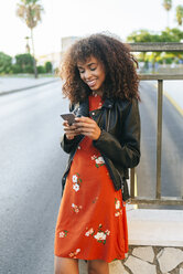Porträt einer lächelnden jungen Frau, die am Straßenrand steht und auf ihr Smartphone schaut - KIJF02169