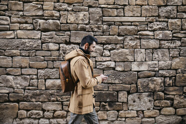 Spanien, Igualada, Mann geht an einer Steinmauer entlang und benutzt ein Mobiltelefon - JRFF02285