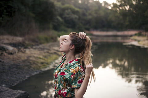 Junge Frau berührt ihr Haar neben einem See, lizenzfreies Stockfoto