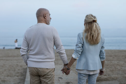 Spanien, Barcelona, Rückansicht eines älteren Paares, das Hand in Hand am Strand in der Abenddämmerung spazieren geht - MAUF02255