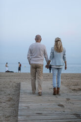 Spanien, Barcelona, Rückansicht eines älteren Paares, das Hand in Hand am Strand in der Abenddämmerung spazieren geht - MAUF02256