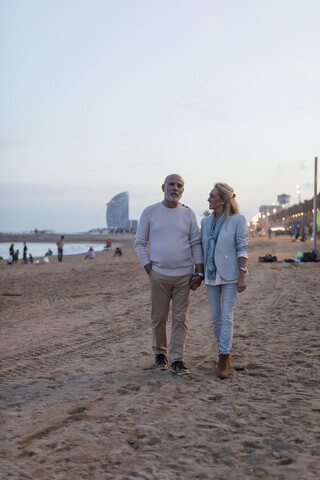 Spanien, Barcelona, älteres Paar, das Hand in Hand in der Abenddämmerung am Strand spazieren geht, lizenzfreies Stockfoto