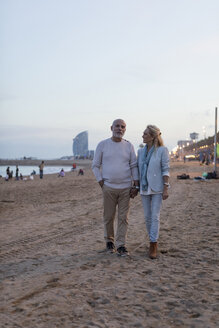 Spanien, Barcelona, älteres Paar, das Hand in Hand in der Abenddämmerung am Strand spazieren geht - MAUF02262