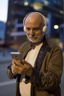 Spanien, Barcelona, älterer Mann mit Kopfhörern und Mobiltelefon in der Stadt in der Abenddämmerung - MAUF02270