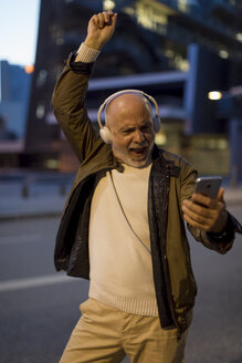 Spanien, Barcelona, aufgeregter älterer Mann mit Kopfhörern und Mobiltelefon in der Stadt in der Abenddämmerung - MAUF02271