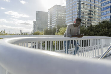 Young man standing on a bridge, listening music, using smartphone - GIOF05354