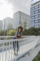 Young woman standing on a bridge, using smartphone, with headphones around her neck - GIOF05356