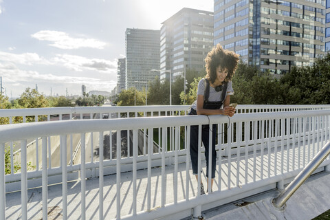 Junge Frau, die auf einer Brücke steht und ein Smartphone benutzt, mit Kopfhörern um den Hals, lizenzfreies Stockfoto