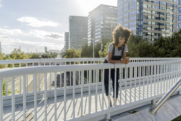 Junge Frau, die auf einer Brücke steht und ein Smartphone benutzt, mit Kopfhörern um den Hals - GIOF05357