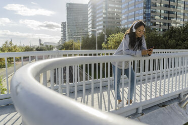 Junge Frau steht auf einer Brücke, hört Musik und benutzt ein Smartphone - GIOF05359