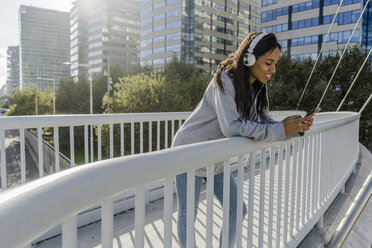 Young woman standing on a bridge, listening music, using smartphone - GIOF05360