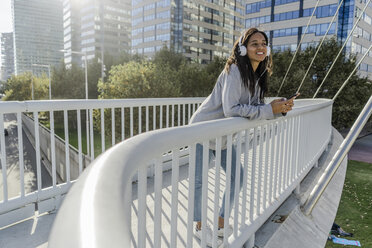 Junge Frau steht auf einer Brücke, hört Musik und benutzt ein Smartphone - GIOF05361