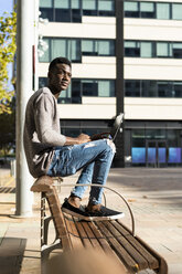 Junger Mann sitzt auf einer Bank in der Stadt und benutzt einen Laptop - GIOF05384