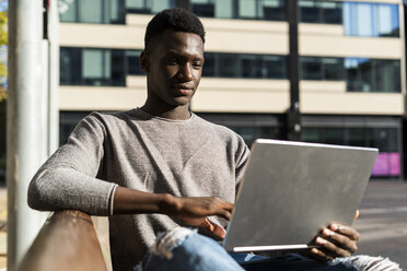Junger Mann sitzt auf einer Bank in der Stadt und benutzt einen Laptop - GIOF05385