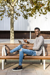 Junger Mann sitzt auf einer Bank in der Stadt und benutzt Laptop und Smartphone - GIOF05388