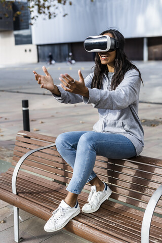 Junge Frau sitzt auf einer Bank, benutzt eine VR-Brille und streckt ihre Hände aus, lizenzfreies Stockfoto