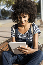 Mid adult woman with curly hair, sitting on a bench, using digital tablet - GIOF05393