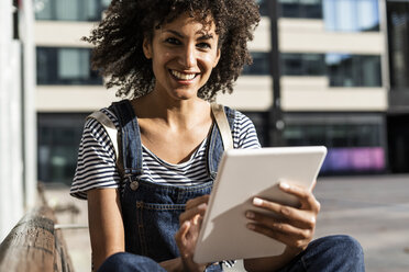 Mid adult woman with curly hair, sitting on a bench, using digital tablet - GIOF05394