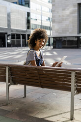 Mittlere erwachsene Frau mit lockigem Haar, auf einer Bank sitzend, mit digitalem Tablet - GIOF05397
