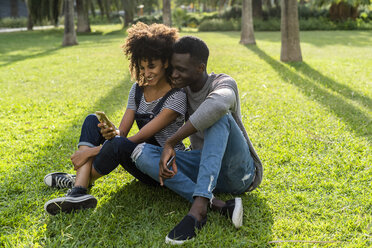 Happy couple sitting on lawn in a park, using smartphone - GIOF05411