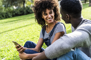 Glückliches Paar sitzt auf einer Wiese in einem Park und benutzt ein Smartphone - GIOF05417