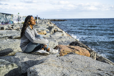 Junge Frau entspannt sich am Strand, auf Felsen sitzend, mit geschlossenen Augen - GIOF05447