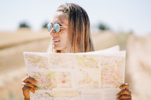 Blonde junge Frau mit verspiegelter Sonnenbrille, die eine Landkarte hält und in die Ferne schaut - OCMF00199