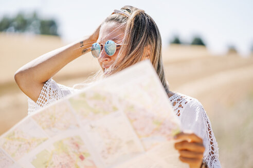 Blonde junge Frau mit verspiegelter Sonnenbrille, die eine Landkarte hält und in die Ferne schaut - OCMF00200