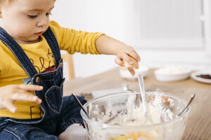 Toddler girl sitting on kitchen table throwing egg into bowl for preparing dough, partial view - JRFF02337