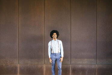 Portrait of smiling man wearing suspenders standing in front of rusty background - JRFF02394