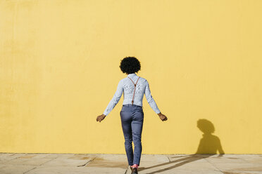 Rückenansicht eines Mannes mit Hosenträgern, der vor einer gelben Wand tanzt - JRFF02412