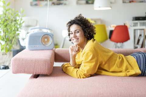 Porträt einer glücklichen Frau, die auf der Couch liegt und mit einem tragbaren Radio zu Hause Musik hört, lizenzfreies Stockfoto