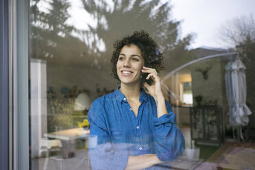 Portrait of smiling woman on cell phone behind window at home - JOSF02735