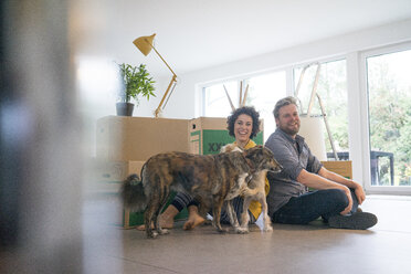Happy couple sitting in living room with dog and cardboard boxes - JOSF02737
