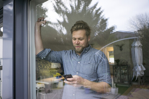 Lächelnder Mann, der zu Hause hinter dem Fenster sein Handy überprüft, lizenzfreies Stockfoto