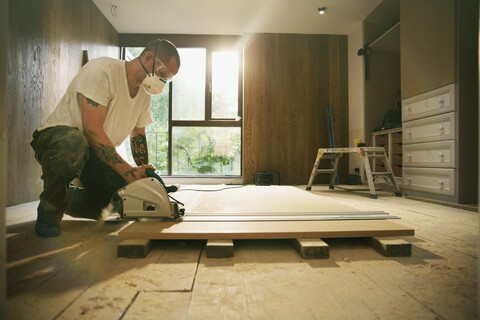 Bauarbeiter mit Tattoos, der eine Tischsäge benutzt, um eine Holzplatte im Haus zu schneiden, lizenzfreies Stockfoto