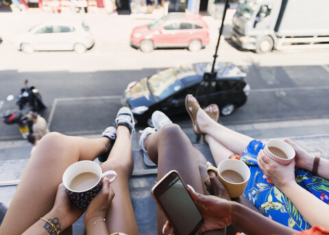 Freundinnen, die Kaffee trinken und die Beine aus dem Fenster einer städtischen Wohnung baumeln lassen, lizenzfreies Stockfoto