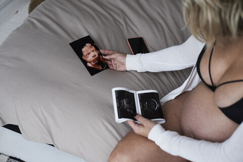 Schwangere Frau schaut sich Ultraschallbilder zu Hause auf dem Bett sitzend an, lizenzfreies Stockfoto