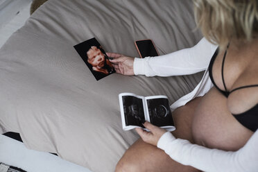 Schwangere Frau schaut sich Ultraschallbilder zu Hause auf dem Bett sitzend an - IGGF00717
