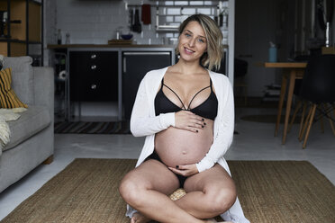 Porträt einer lächelnden blonden schwangeren Frau, die zu Hause auf dem Boden sitzt - IGGF00726