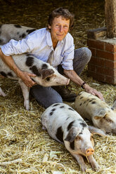 Lächelnde ältere Frau kniend in einer Scheune mit Gloucester Old Spot Schweinen. - MINF09838