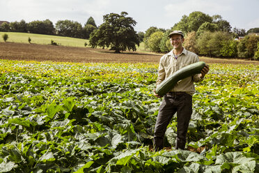 Ein lächelnder Bauer steht auf einem Feld und hält einen großen grünen Kürbis. - MINF09856